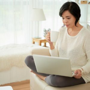 穿白色長袖灰色褲子拿著咖啡看著電腦影片的女生